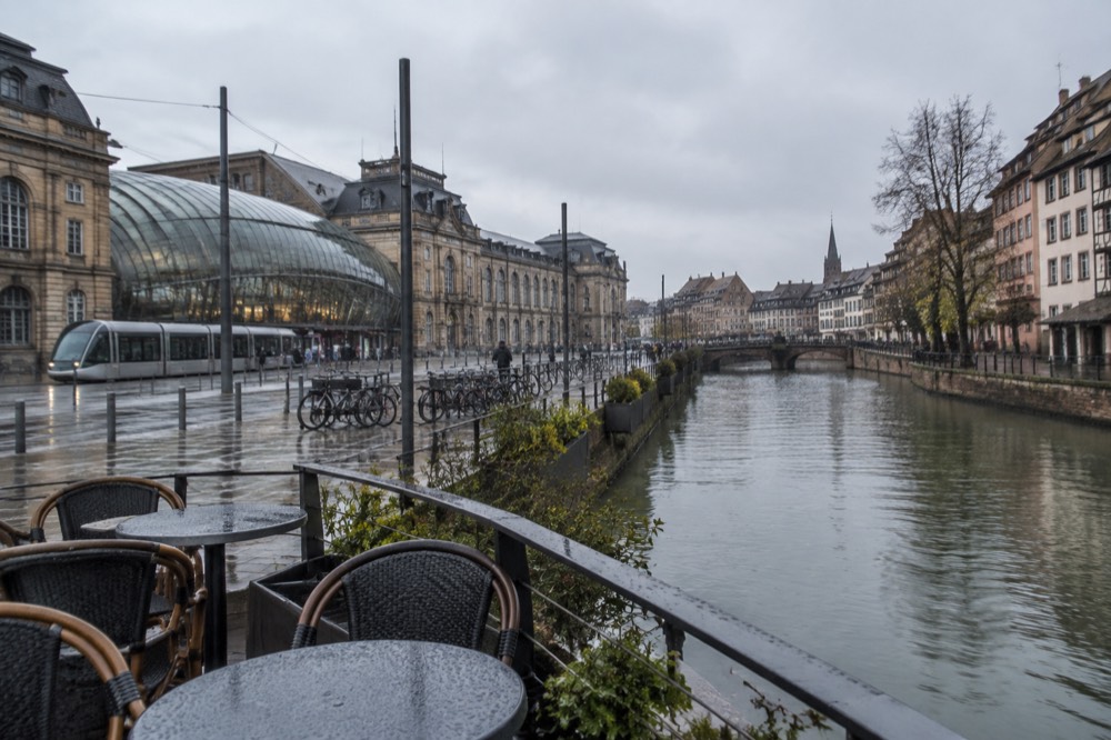 A riverside terrace with chairs overlooking canal waters reflecting old buildings