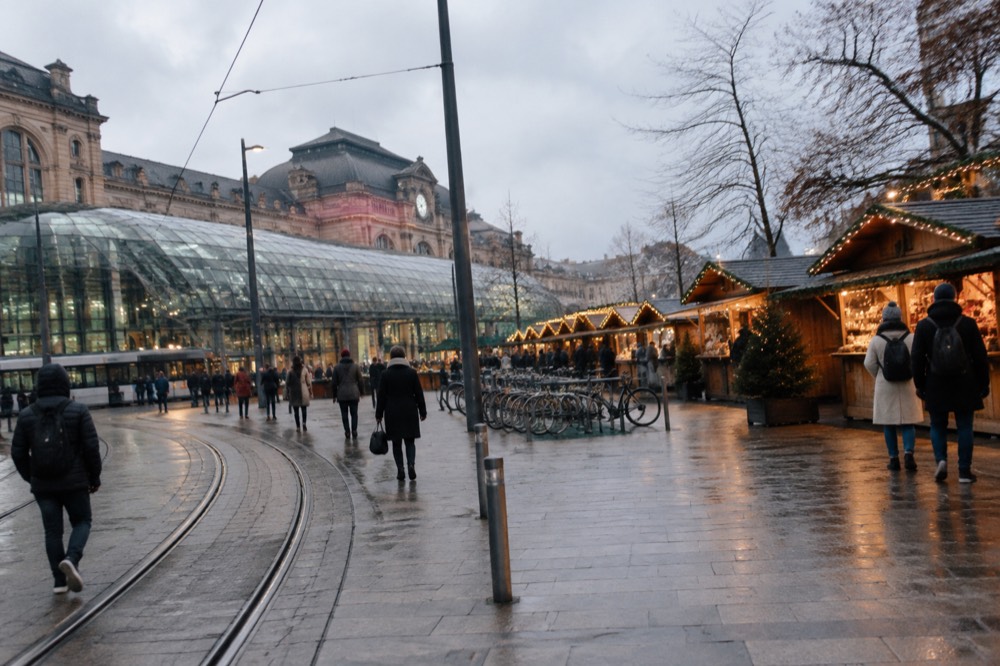 A Christmas market square at dusk with wooden chalets glowing under strings of lights
