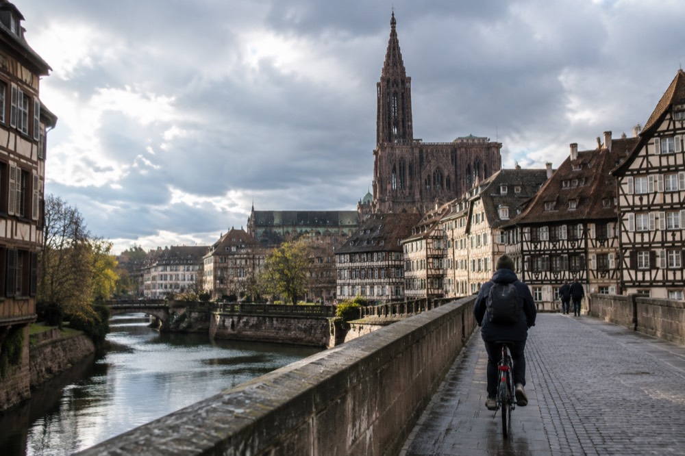 A cyclist crossing a stone bridge over the Ill with the cathedral spire in the distance