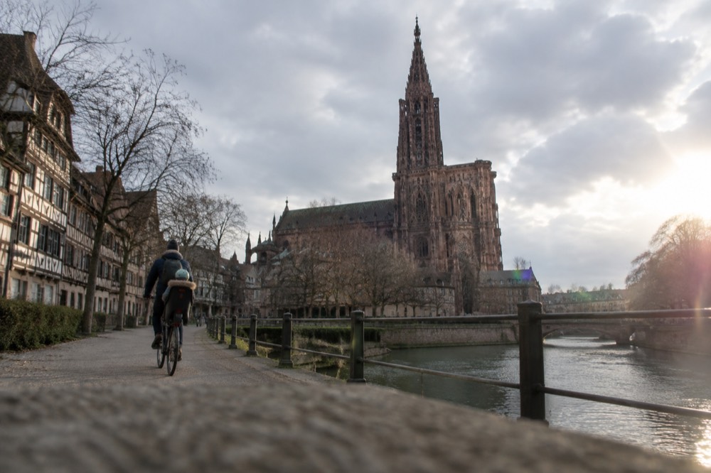 A parent cycling with a child on a bike seat along a wide riverside path