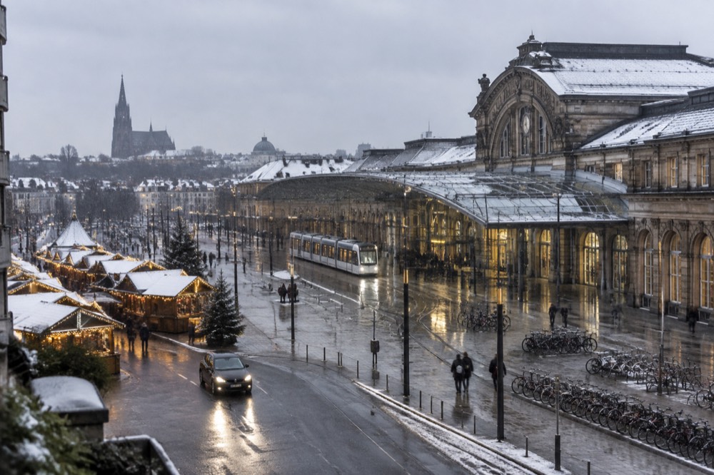 A car with headlights on passing illuminated market chalets in a snowy European square