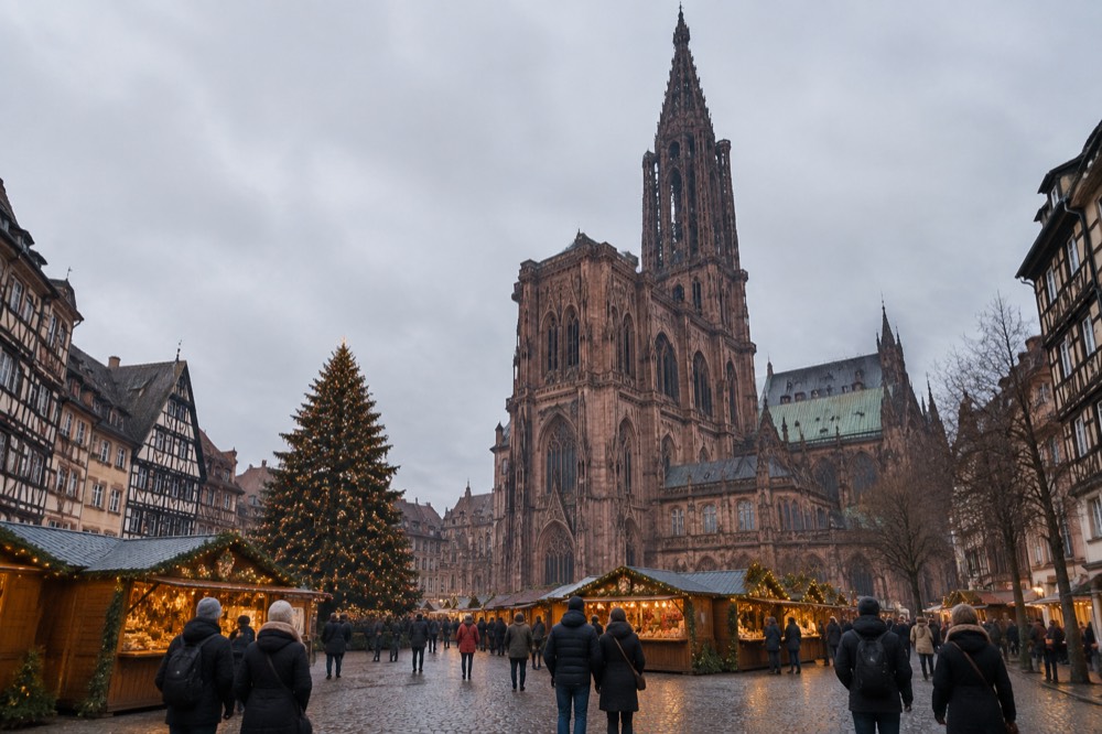 Illuminated Christmas market chalets and a large decorated tree beside a Gothic cathedral at dusk