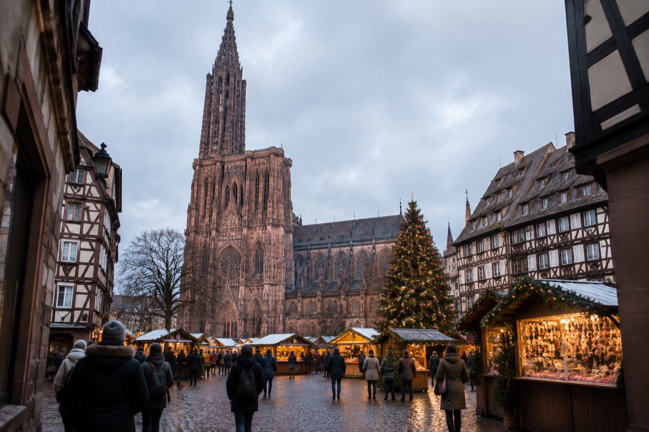 Illuminated Christmas market chalets and a large decorated tree beside a Gothic cathedral at dusk