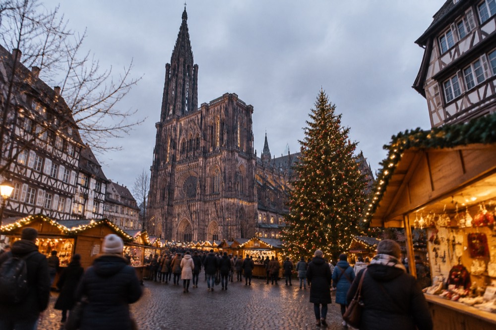 A Christmas market square in Strasbourg with timber chalets and a large decorated tree at dusk