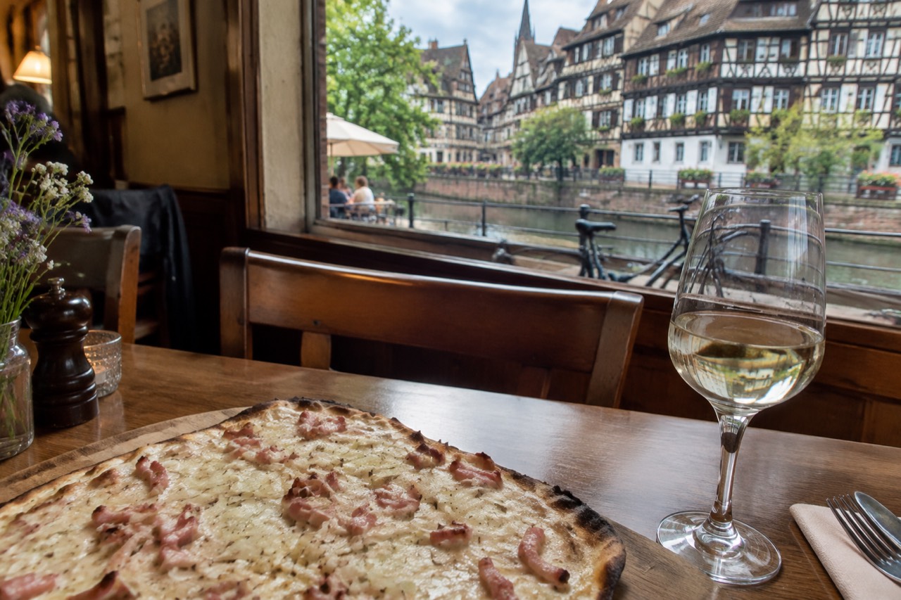 A plate of tarte flambée and a glass of white wine on a wooden table in a warm Alsatian dining room