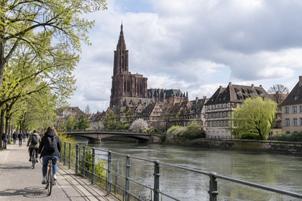 Cyclists on a riverside path in Strasbourg with the cathedral visible beyond the rooftops