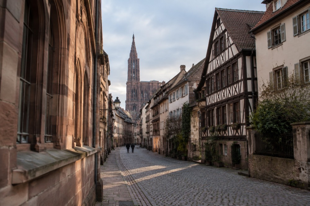 A narrow medieval street with sandstone buildings leading toward Strasbourg Cathedral's single spire