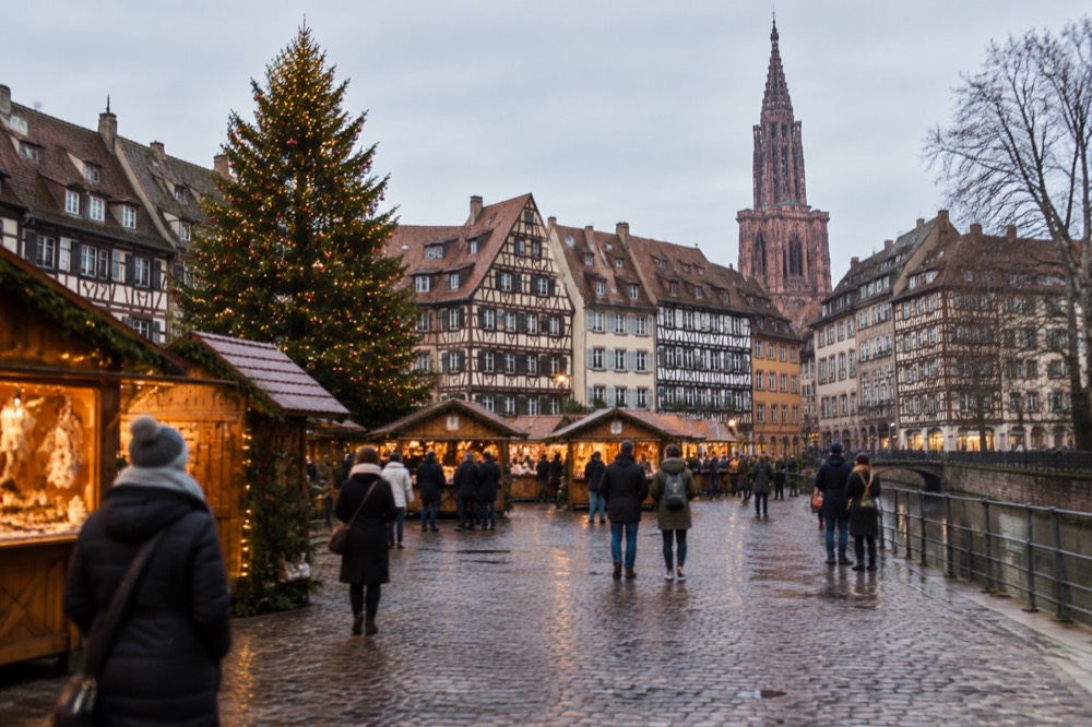 A Christmas market square at dusk with a large decorated tree and wooden chalets