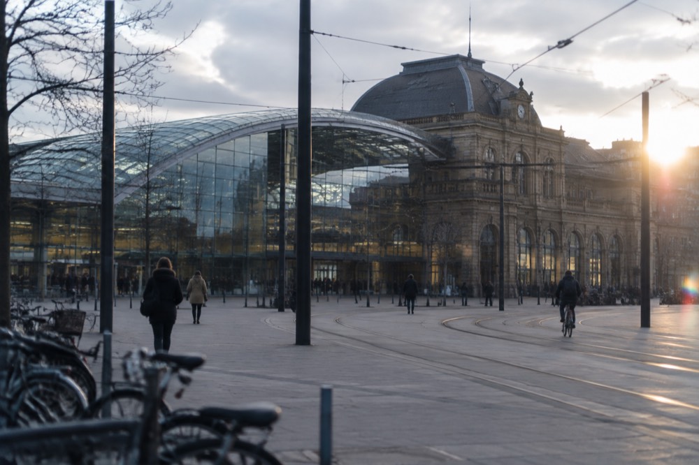 The glass and steel frontage of a large railway station with tram tracks running along the plaza outside