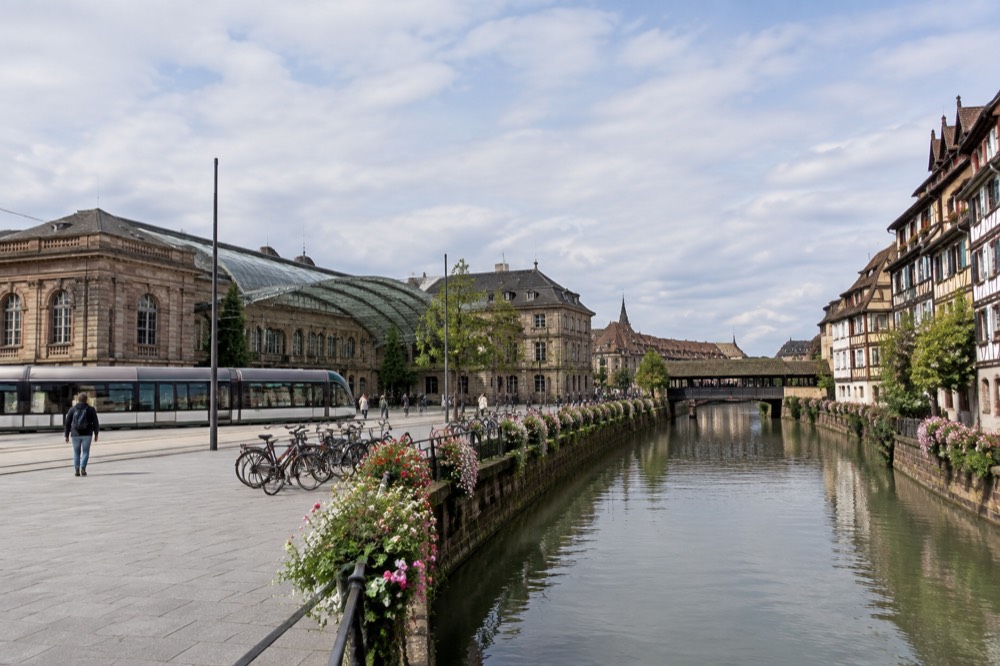A canal lined with flowers and half-timbered buildings leading towards a covered bridge