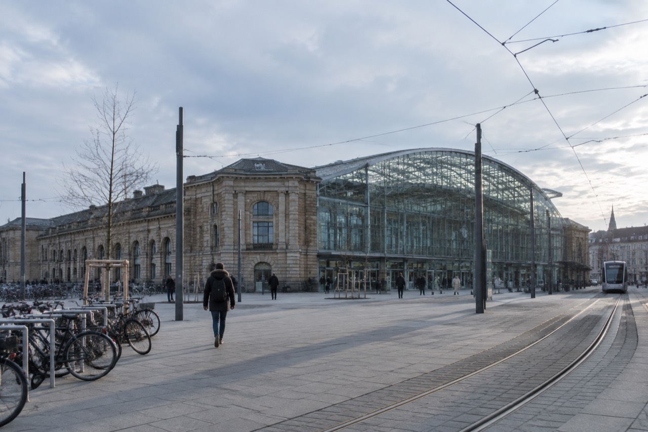 The glass and steel frontage of a large railway station with tram tracks running along the plaza outside
