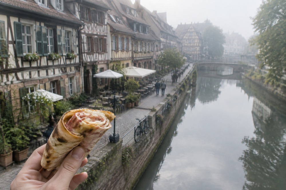 A rolled slice of tarte flambée held between fingers, showing the thin fold of crust