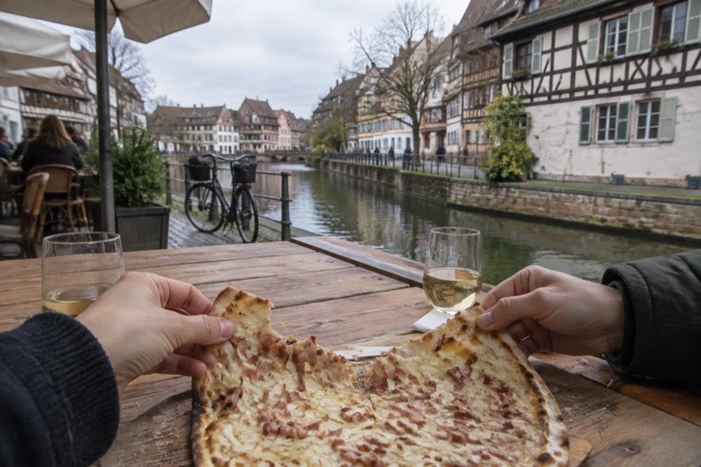 Hands tearing a shared tarte flambée apart at a long wooden table