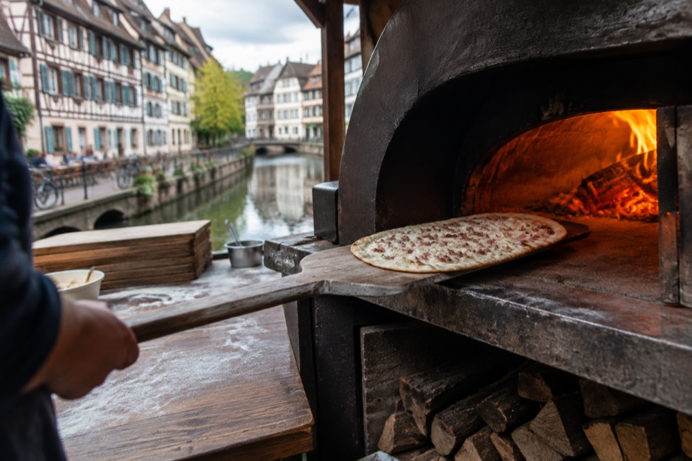 A tarte flambée being slid from a wooden peel into the glowing mouth of a wood-fired oven