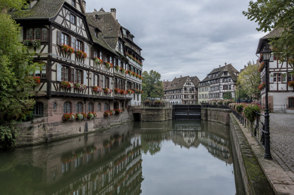 Flower-decked half-timbered buildings reflected in a still canal in Petite France