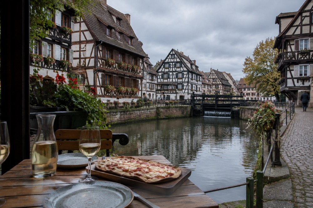 A wooden winstub table set with stoneware plates, a carafe of white wine and a board of tarte flambee