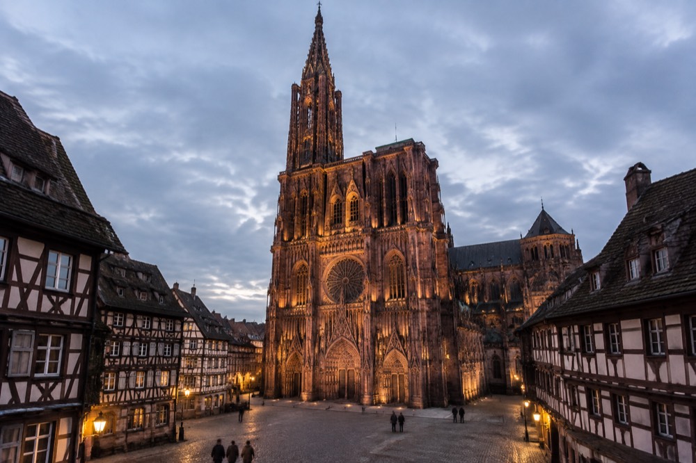 The floodlit west facade of Strasbourg Cathedral seen at dusk from a cobbled square