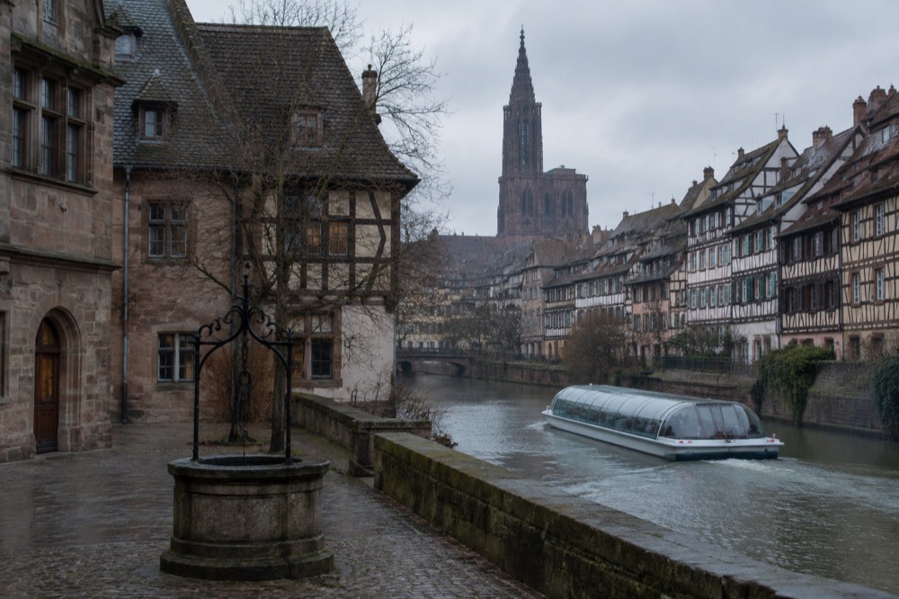 Covered river boat passing half-timbered houses along the Ill in light rain